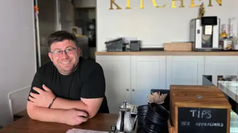 Jonny Humphries/BBC Emil Rahaeanu, wearing a black t-shirt and glasses, smiles at the camera from behind the counter at his café