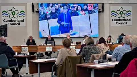 Dozens of councillors are gathered for a meeting in a white room with wooden desks and signs on the wall reading: "Cumberland Council". A large screen behind the head table shows Josh Kirkwood standing and reading from a piece of paper. There are dozens of people sitting in the public gallery behind him.