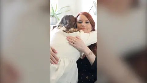 Woman smiles as she hugs a woman wearing a cream dress.