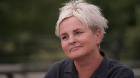 Sue Davison has short blonde hair and brown eyes. She wears a black shirt and is looking away from the camera, listening to the interviewer
