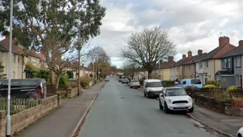 Google A long residential street with houses either side. There are cars parked on both sides of the road and large trees next to houses that are bare or have little green leaves