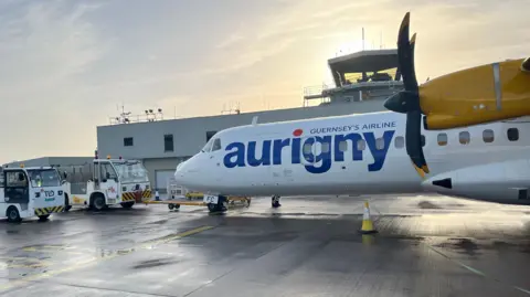 Image of Aurigny propeller plane parked at the airport on the right hand side. The sun is glowing behind the watchtower. Two airport vehicles are parked to the left of the plane. 