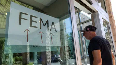 Reuters An elderly man wearing a baseball cap enters an office with a large sign saying FEMA and three red arrows 