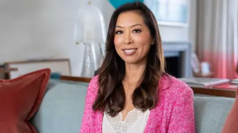 Vivien Wong wearing a white lace camisole and bright pink wool cardigan sitting on a grey sofa in a plush home