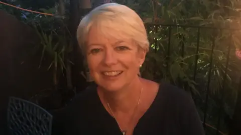 Lorraine Lorraine smiling wearing a gold chain with cropped blonde hair and a dark blue top.