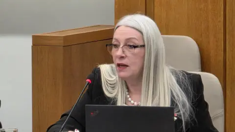 Kay Mason Billig is sitting in the Norfolk County Council chamber. She is wearing glasses, a white pearl neckless and black suit jacket. In front of her is a laptop and microphone.