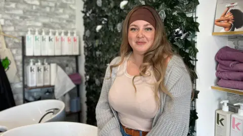 BBC Sophie Reid sitting in a salon or spa setting. In the background, there are shelves stocked with hair care products, neatly folded towels, and a decorative wall featuring floral elements. In the foreground, two white sinks typically used for hair washing are visible.