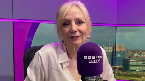 A woman with scraped-back blond hair wearing a cream blouse and sitting in a radio studio with a purple background