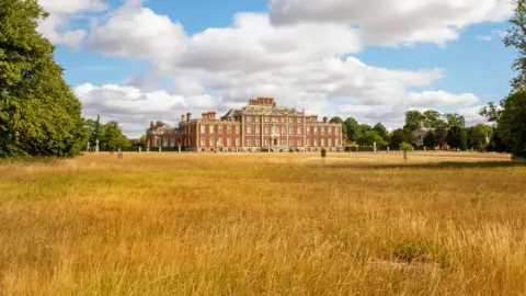 A large stately home is in the distance. A large field which has dry grass and hay leads to the home which is three storeys. Large green trees are surrounding the building.