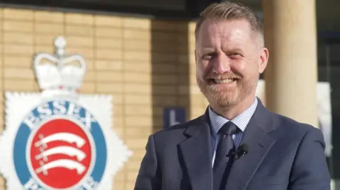 Jamie Niblock/BBC DS Gary Biddle wears a blue suit and tie, and stands outside a building with an Essex Police crest.