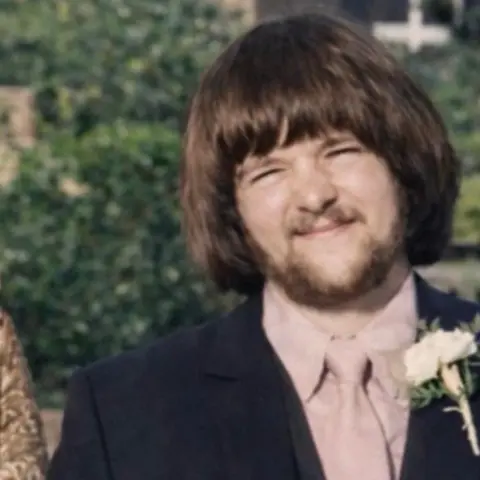 Family handout An archive image of Graham Finn with short brown hair and a fringe reaching his eyebrows. He is wearing a dark suit with a pink shirt and pink tie, and a white carnation pinned to his blazer lapel. He is standing in front of a green bush.