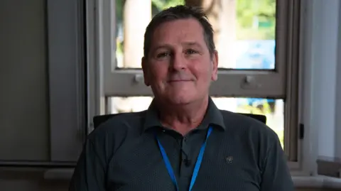 Russell seated indoors in front of a window, wearing a dark collared shirt and blue lanyard.
Russell is clean shaven and has short, spikey hair.