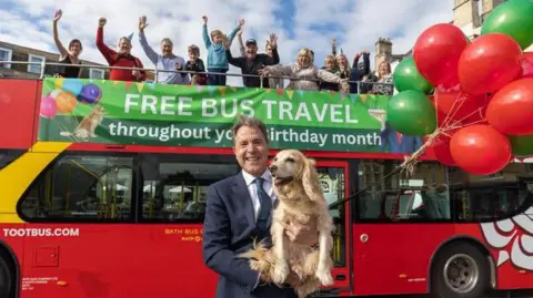 West of England Mayor Dan Norris holding his dog, with a red bus behind him with children on the top deck waving at the camera