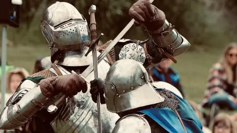 Jersey Heritage Two people in silver armour and chainmail are battling eachother and have their swords crossed. The one with their back to the camera is also wearing a blue silky cape. They have full visors and are wearing brown leather gauntlets. Behind them, out of focus, are spectators.
