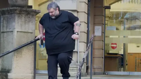 A picture of Barry O'Sullivan walking down the steps at Reading Crown Court with a crutch in his left hand and book in his right hand.