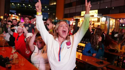 PA Media Football fans at the EV Charge Points Stadium in Cheltenham throw their hands up in the air in excitement as England win their first World Cup game.