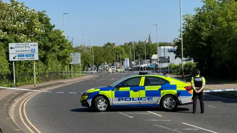 A police cordon in Welford Road, Leicester. One police car is parked in the middle of the road with an officer stood outside, facing away from the camera. Police tape can be seen across the road in the distance