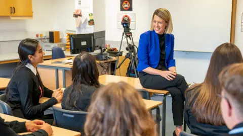 Ministra vzdělání PA Jenny Gilruth během návštěvy Stonelaw High School v Rutherglenu. Sedí na stole a mluví se žáky.