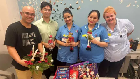 North West Anglia Hospitals' Charity Five people are stood next to each other. They are all looking at the camera and the four on the left are holding different Christmas items. 
