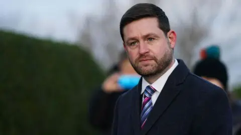 A man in a dark coat, white shirt and multicoloured striped tie stands in a suburban street. There is a hedge behind him and some people out of focus. 