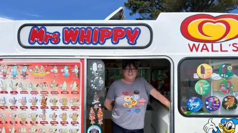 Sarah McKeich stood inside an ice cream van. She is smiling at the camera, and is wearing a striped top with badges on it. There are pictures of ice creams on the windows.