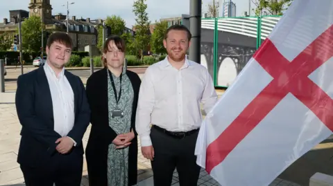 Three Reform councillors - two men and a woman - on Sunderland City Council are stood outside the City Hall. One is holding a St George's Flag which has been raised. They are stood in the shade, and behind them, the street is lit by warm sunlight. They are all dressed in formal attire.