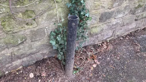 LDRS The stump of a post box. The post box is gone. All is left is the black metal stump of the box. The stump is on a pavement, in front of a stone wall.