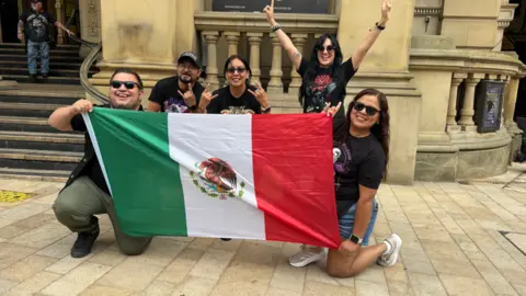 A group of two men and three women hold up a Mexican flag outside Birmingham Town hall and grin at the camera.