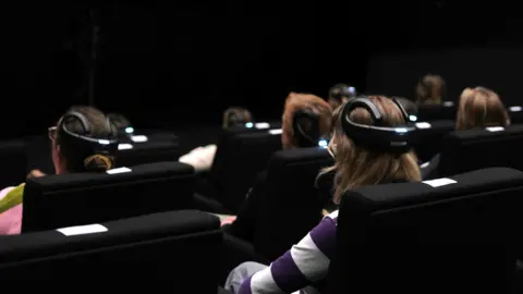 Audience members sitting in an auditorium. They are wearing head sets and looking at a dark screen.