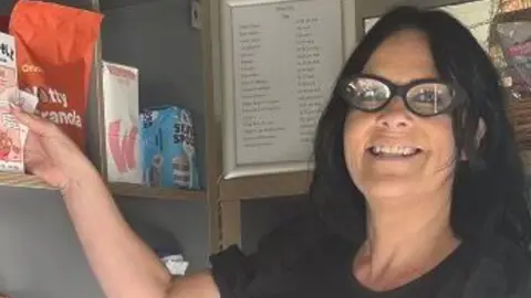 Rachael Parke is standing in her community shop in front of shelves of food. She has shoulder length black hair, wears black glasses and is holding a mug of coffee and a box of cereal.