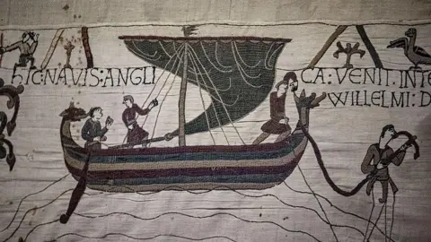 Getty Images Detail of the Bayeux Tapestry which shows figures in and around a boat with wording telling the story embroidered alongside it 