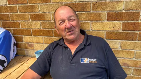 Kate Bradbrook/BBC A man sits at an outdoor pub table and smiles at the camera. He is largely bald and is wearing a navy polo shirt. He is leaning against a yellow brick wall.
