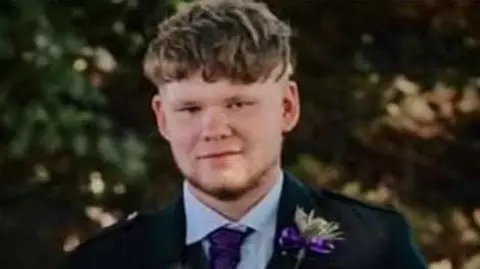 Calum Carmichael looking straight at the camera. He is wearing a blue suit jacket with a white shirt and purple tie. He has a purple flower in his lapel pin.