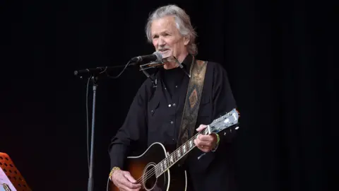 Kris Kristofferson performing on the Pyramid stage at Glastonbury 2017. He is singing into a microphone, is strumming a brown and white guitar and has a harmonica around his neck. The background is totally black.