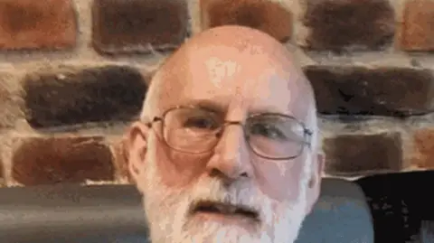 Roddy MacPherson, a man with glasses, balding with white hair and a white beard smiles into the camera while sitting indoors, in front of a brick wall.