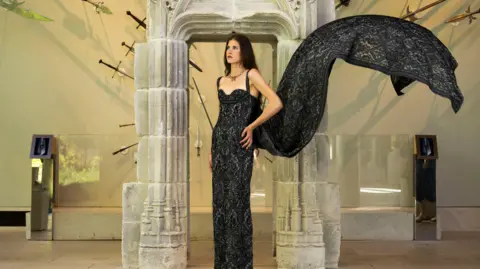 Danae Crawford - a woman with long dark hair, wearing a flowing black dress, standing in a museum with antiques around her 