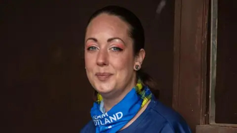 Sheree Gregg smiles at the camera. She has her dark brown hair tied back and wears pink eyeshadow. She has a blue and yellow Team Scotland scarf and wears a blue -tshirt.