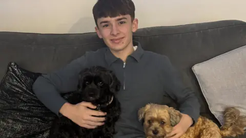 Finley has short dark hair and wears a dark zip-up top. He is sat on a sofa with the family's two lassapoos.