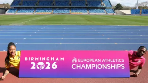 Birmingham European Athletics Championships 2026 Amber Anning is wearing a yellow T-shirt and has long braided hair she is lying behind a pink and purple sign with the logo of Birmingham 2026 on. At the other end Jeremiah Azu lies behind the sign and is wearing a pink tee-shirt