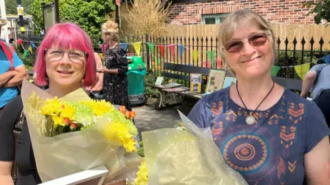Two women stand on the station platform , they have both been presented with a bouquet of flowers.
