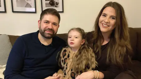 Sitting on the sofa at home are Alba and her parents Dan and Melissa