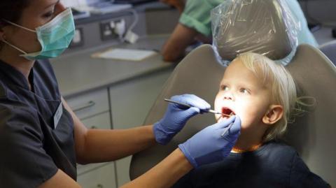  A young boy sits in the dentist chair as a nurse with blue gloves on pokes around in his mouth with tools.