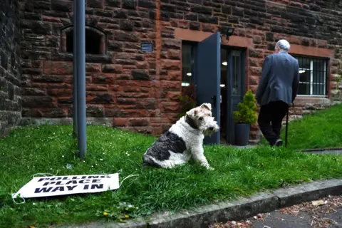 Andrew Milligan / PA Media The terrier has black and white fur and sits next to a sign lying on the ground saying 'polling place way in' while a man walks into the station with a cane in hand.