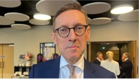 Ian Gorst pictured at a finance event. He is wearing a blue suit, and a white shirt. He is also wearing glasses. 