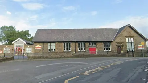 A single story school building with a red door and several windows. It has a slate roof. There is a playground in front and railings around it.