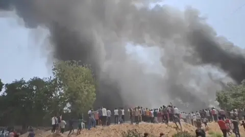 A group of people stand near a plane crash site, as black smoke billows into the air.
