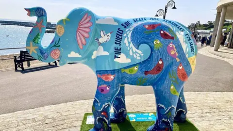 A fibreglass elephant stands on a platform next to a beach. It appears to be about twice the height of a nearby bench. It is painted mainly in blue, with fish and sea creatures.