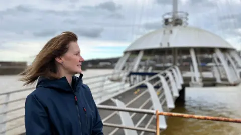 Deanne Shallcross, CEO of Whitehaven Harbour Commissioners. She is stood at Whitehaven Harbour by the walkway that leads to the Crow's Nest - a pagoda-type white structure on the water. She is looking out at the harbour, side-on with the wind blowing back her shoulder-length brown hair. She is wearing a blue waterproof jacket. The background is blurred.