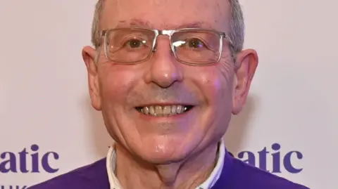 Rachel Smith A man in a purple jumper wearing rectangular glasses smiles at the camera