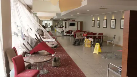 Interior of a hotel dining area, with chairs having been thrown through a window and broken glass scattered across seats, tables and the floor